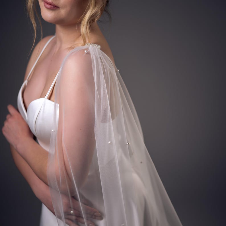 May include: A sheer white tulle capelet featuring scattered small white pearls attached to the fabric. The capelet is draped over the shoulders of a white satin dress with thin spaghetti straps, creating a soft, layered appearance.