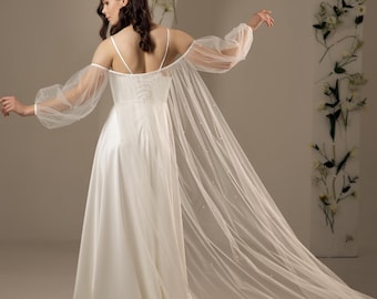 May include: A white bridal gown with delicate spaghetti straps and a flowing skirt. The dress has sheer, long sleeves embellished with tiny pearls. A long, sheer veil with pearl detailing trails behind the dress, creating an elegant silhouette.