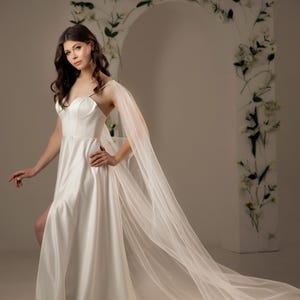 May include: A floor-length white satin gown featuring a sweetheart neckline, thin spaghetti straps, and a high side slit. The dress is paired with long, sheer, flowing tulle cape sleeves that extend to the floor, creating a dramatic train effect.