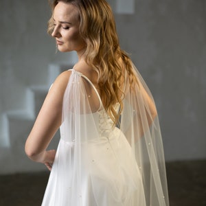 May include: A white floor-length dress featuring a corset-style bodice with thin straps and a sheer tulle cape attached at the shoulders. The cape is adorned with small, evenly spaced white pearls and flows down the back, creating a soft, layered effect.