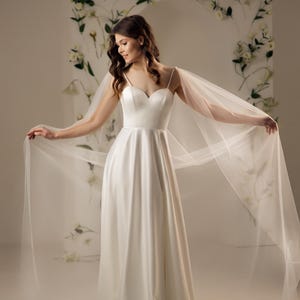 May include: A floor-length white satin gown featuring a sweetheart neckline, thin spaghetti straps, and a full skirt. The dress includes long, sheer, flowing tulle cape sleeves that extend from the shoulders, creating a dramatic, airy silhouette.