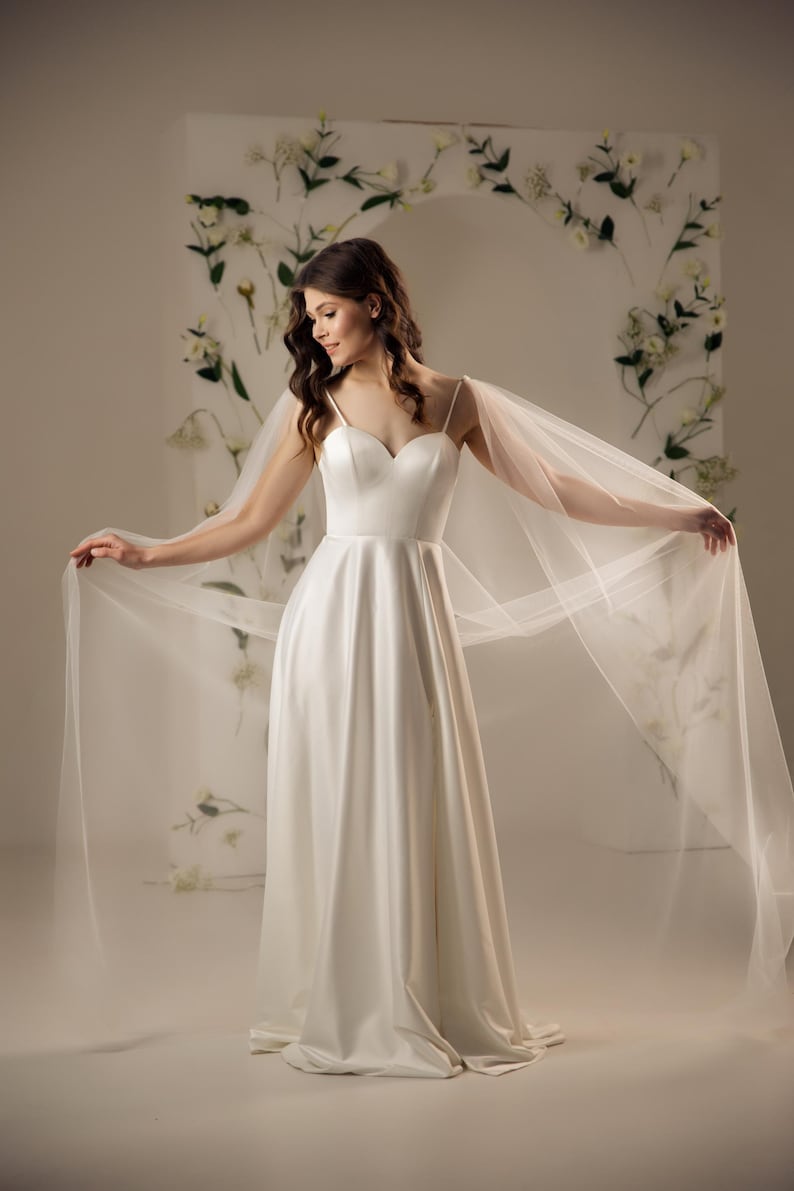 May include: A floor-length white satin gown featuring a sweetheart neckline, thin spaghetti straps, and a full skirt. The dress includes long, sheer, flowing tulle cape sleeves that extend from the shoulders, creating a dramatic, airy silhouette.