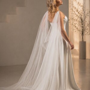 May include: A floor-length white gown featuring a smooth, solid bodice with thin straps and a flowing skirt. Attached to the shoulders are long, sheer, translucent tulle panels that drape down the back, creating a sweeping train effect.