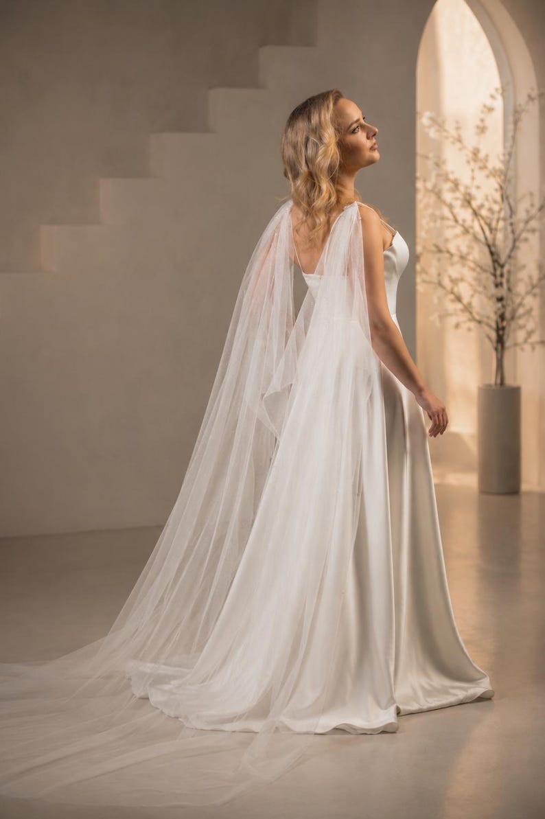 May include: A floor-length white gown featuring a smooth, solid bodice with thin straps and a flowing skirt. Attached to the shoulders are long, sheer, translucent tulle panels that drape down the back, creating a sweeping train effect.