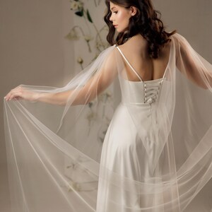 May include: A floor-length white satin dress featuring thin spaghetti straps and a lace-up corset back closure. The garment includes sheer, flowing tulle cape sleeves that extend from the shoulders, creating a dramatic, airy silhouette.