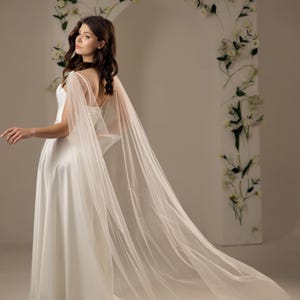 May include: A floor-length white bridal gown featuring a smooth, solid fabric bodice and a long, flowing sheer tulle cape attached at the shoulders. The cape is adorned with small, scattered white pearls and extends behind the dress.