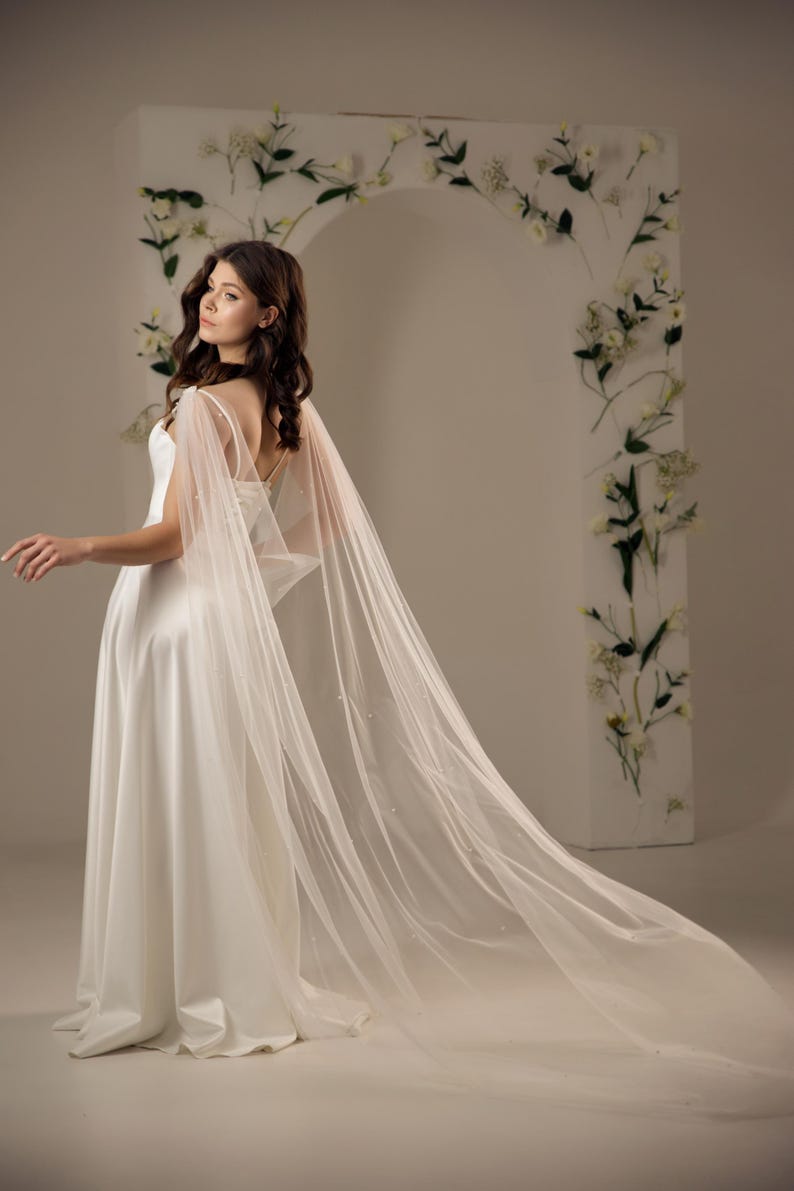 May include: A floor-length white bridal gown featuring a smooth, solid fabric bodice and a long, flowing sheer tulle cape attached at the shoulders. The cape is adorned with small, scattered white pearls and extends behind the dress.