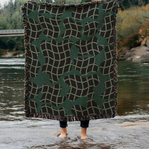 May include: A woven throw blanket with a dark green and black grid pattern. The blanket has a fringed edge and is held up by a person standing in a river. The pattern is a repeating design of curved lines.