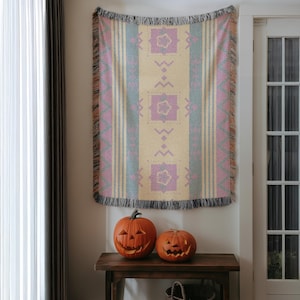 May include: A woven tapestry with a cream-coloured central panel, adorned with lavender floral and geometric patterns, flanked by vertical stripes of lavender and teal. The tapestry is fringed at the top and bottom. Two carved pumpkins sit on a wooden table.