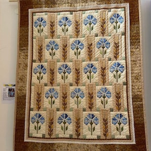 May include: A quilt featuring a repeating pattern of blue flowers and wheat stalks in shades of brown, beige, and green. The quilt is framed with a brown border and white lace.