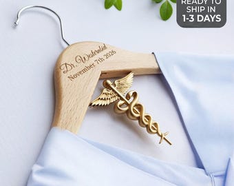 May include: A light-coloured wooden clothes hanger featuring a gold-toned metal caduceus emblem attached to the centre. The wood is engraved with the text Dr. Westernbid and the date November 7th 2026. A light blue garment is draped over the hanger.