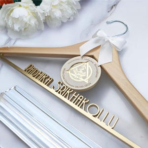May include: A wooden hanger with a gold inscription that reads "Radhika Srihakolu" and a gold University of Alabama at Birmingham White Coat Ceremony medallion with the date 08/09/2024.