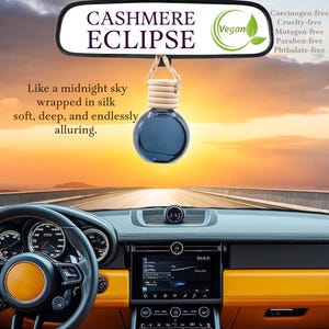 May include: A car air freshener with a black glass bottle and a wooden cap. The bottle is hanging from a rearview mirror with the text "CASHMERE ECLIPSE" and "Vegan" on the mirror. The text "Like a midnight sky wrapped in silk soft, deep, and endlessly alluring." is below the bottle. The background is a sunset sky.