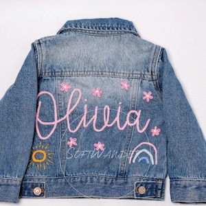 May include: A blue denim jacket with pink embroidery of the name "Olivia" and a sun, flowers, and rainbow design.