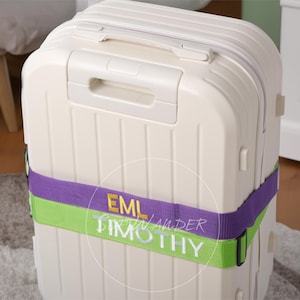 Puede incluir: Una maleta rígida de color crema con ranuras verticales y un asa superior. Una correa de equipaje de color morado y verde lima está envuelta horizontalmente alrededor del centro, bordada con el texto amarillo EML y el texto blanco TIMOTHY.