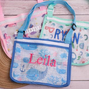 Peut inclure: Trois sacs de plage en filet avec des bretelles réglables. L'un est rose avec un motif de licorne, l'autre est bleu avec un motif de baleine et le nom "Leila", et l'autre est vert avec le nom "Ryan". Les sacs sont sur une surface en bois.