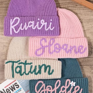 May include: Four knitted beanies in different colors with names embroidered on them. The beanies are stacked on top of each other. The names are Ruairi, Sloane, Tatum, and Goldie.