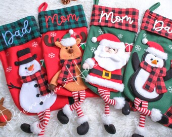 May include: Four Christmas stockings with festive designs. One red stocking has a snowman, another has a reindeer, and a third has Santa Claus. The fourth stocking has a penguin. Each stocking has a plaid cuff and the names "Dad" and "Mom".