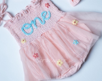 May include: A pink tulle baby dress with embroidered flowers and the word "one" in blue thread. The dress has thin straps and a ruffled neckline.