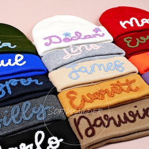 Peut inclure: Une pile de bonnets en tricot colorés avec des noms brodés, dont Ella Paul,  Faspree, Billy, Carlo, Declan, Lina, James, Everett, Darwin,  Evelyn,  Felix, Mia, et  H.