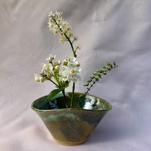 Op de afbeelding: Een keramische kom met een groene en bruine glazuurlaag bevat een bloemstuk. Witte bloemen en groene stelen zijn in de kom gerangschikt. De kom heeft een decoratief patroon aan de zijkant.