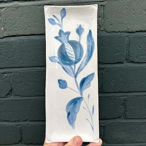 May include: A rectangular white ceramic serving dish with a hand-painted blue floral design. The design features a central pomegranate-like flower with detailed petals and leaves. The dish is held against a dark green brick wall.