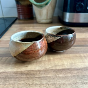 Può includere: Due tazze in ceramica fatte a mano con un liquido scuro all'interno. Le tazze hanno una forma arrotondata con un mix di colori marrone, beige e arancione. Le tazze sono su una superficie di legno. Ogni tazza misura circa 8 cm di diametro.