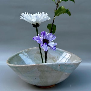 Op de afbeelding: Een keramische kom met een lichtblauwe en witte glazuur bevat een witte en een paarse chrysant. De kom heeft een unieke, asymmetrische vorm en een roodbruine basis. De bloemen zijn geschikt voor een minimalistische bloemenpresentatie.