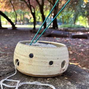 May include: A cream-coloured ceramic yarn bowl with a speckled texture and multiple circular openings along the side. A ball of light grey yarn sits inside the bowl, with a strand threaded through one hole, and two thin green knitting needles.