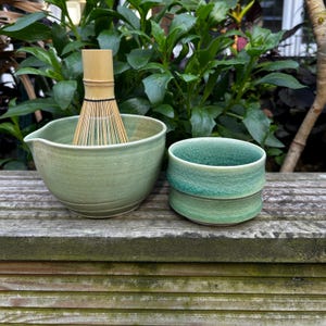 Könnte beinhalten: Ein Matcha-Teeset mit einer hellgrünen Keramikschale mit einem Bambusbesen und einer kleineren, strukturierten grünen Keramiktasse. Das Set wird auf einer verwitterten Holzoberfläche mit üppigem grünem Laub im Hintergrund präsentiert.