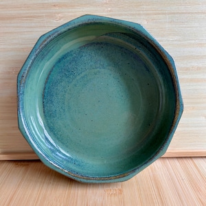 Op de afbeelding: Een handgemaakte, achthoekige keramische kom met een turquoise en blauwe glazuur. De kom heeft een getextureerd oppervlak en een bruine rand. De binnenkant van de kom is een lichtere tint turquoise, waardoor een gradiënteffect ontstaat. De kom staat op een houten oppervlak.