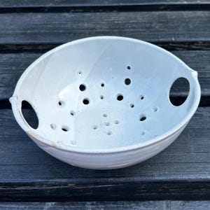 Puede incluir: Un colador de cerámica blanca con numerosos orificios de drenaje. El cuenco tiene dos asas grandes y redondeadas y una superficie ligeramente texturizada. El colador está diseñado para escurrir líquidos de los alimentos.