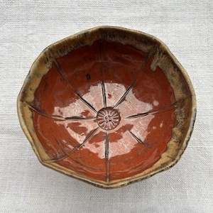 Peut inclure: Un bol en céramique fait à la main avec un design géométrique unique. Le bol présente un intérieur rouge-orangé profond et un bord brun texturé. L'intérieur est orné d'un motif en étoile rayonnant d'un motif floral central.