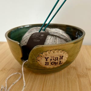 May include: A handcrafted ceramic yarn bowl in green and brown tones, featuring the text "YARN BOWL". It contains a ball of grey yarn and two green knitting needles. A length of yarn trails from the bowl, ideal for knitting or crochet.