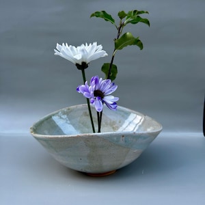 Può includere: Una ciotola in ceramica per ikebana con smalto azzurro e beige, contenente un crisantemo bianco, un crisantemo viola e uno stelo verde e foglioso. La ciotola ha una forma triangolare con angoli arrotondati.