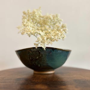 Op de afbeelding: Een handgemaakte keramische kom met een donkergroene glazuur en een lichtbruine rand. De kom is gevuld met een tros kleine, witte bloemen. De kom staat op een donker houten oppervlak.
