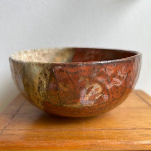 Op de afbeelding: Een handgemaakte keramische kom met een rustieke, getextureerde uitstraling. De kom heeft een mix van aardetinten, waaronder bruin, oranje en beige. De binnen- en buitenoppervlakken vertonen unieke variaties in kleur en textuur.