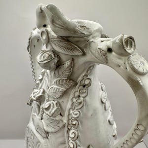 Op de afbeelding: Witte keramische kan met ingewikkelde bloemen- en vogeldetails. De kan heeft verhoogde rozen, bladeren en een decoratieve rand. De handgreep is gebogen en versierd met cirkelvormige patronen. Een vogelsculptuur zit op de tuit.