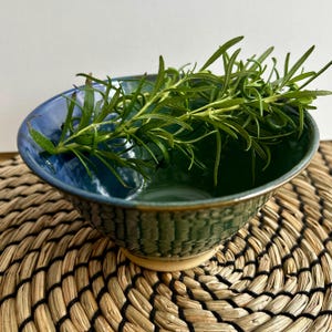 Peut inclure: Un bol en céramique fait à la main avec un extérieur texturé et un intérieur brillant bleu et vert. Des brins de romarin frais reposent à l'intérieur du bol. Le bol est posé sur un dessous de plat tressé.
