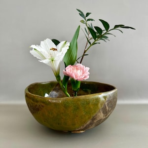 Op de afbeelding: Een keramische ikebana-kom in groen- en bruintinten bevat een bloemstuk. Het arrangement omvat een witte lelie, een roze anjer, groene bladeren en een bladrijke tak. De kom heeft een rustieke, organische vorm, perfect voor het presenteren van bloemen.