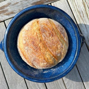 May include: A round loaf of freshly baked bread sits in a deep blue ceramic bowl. The bread has a golden-brown crust with visible scoring. The bowl has a glossy finish and is placed on a wooden surface, creating a rustic, inviting scene.