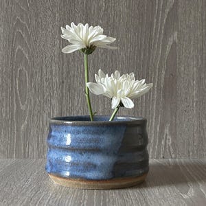 Op de afbeelding: Een kleine, handgemaakte keramische vaas in blauwe en bruine tinten, met twee witte chrysantenbloemen. De vaas heeft een textuur, geribbeld ontwerp en een glanzende afwerking. De bloemen hebben delicate witte bloemblaadjes en groene stelen.