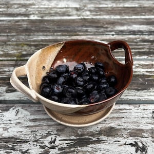 Può includere: Una ciotola in ceramica fatta a mano con una glassa marrone e beige, piena di mirtilli freschi. La ciotola ha un manico su ogni lato e poggia su un piattino abbinato. Il design rustico è posto su una superficie di legno invecchiato.