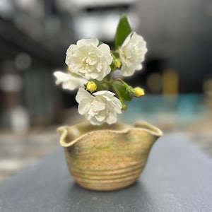 Op de afbeelding: Een kleine, handgemaakte keramische kom met een gegolfde rand, met een takje witte bloemen en groene bladeren. De kom heeft een gespikkelde, aardse kleurenpalet van bruin en groen. De bloemen staan in volle bloei, met gele knoppen.