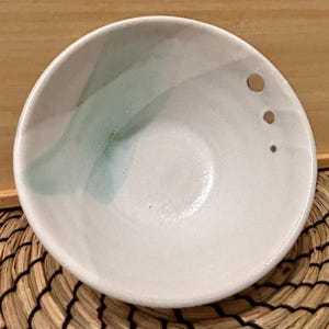 May include: A small, white ceramic bowl with a minimalist design. The bowl has a subtle, abstract teal and grey brushstroke pattern on the interior. Three small, evenly spaced holes are drilled into the rim. The bowl is set on a woven mat.