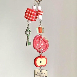 May include: A keychain featuring a silver metal clasp, white and red beads, a red gingham cube, a small green key charm, a red apple-shaped button, a red apple charm, and a silver rectangular pendant with the text follow your dreams trust your heart.