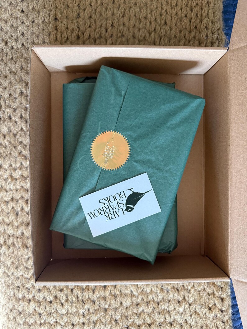 May include: A brown cardboard box with a green tissue paper wrapped package inside. The package has a gold seal and a business card that says "Lark Sparrow Books".