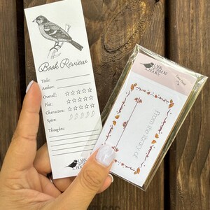 May include: A white bookmark with a black and white illustration of a bird. The bookmark has a section for a book review with headings for title, author, overall, plot, characters, spice, and thoughts. The bookmark also has a section for rating the book with stars and chili peppers.