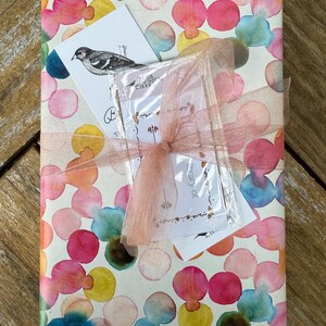 May include: A gift wrapped in white paper with a colorful watercolor pattern of pink, blue, yellow, and green circles. The gift is tied with a pink ribbon and has a small card attached with the text "Birthday of".