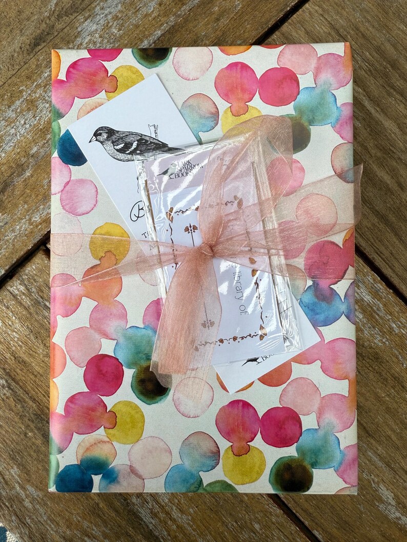 May include: A gift wrapped in white paper with a colorful watercolor pattern of pink, blue, yellow, and green circles. The gift is tied with a pink ribbon and has a small card attached with the text "Birthday of".